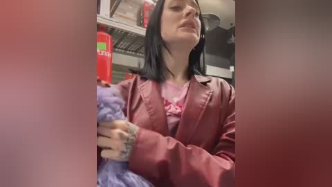 A video of a woman with shoulder-length black hair, wearing a pink satin blouse and a red jacket, cleaning with a purple sponge in a cluttered, dimly lit kitchen.