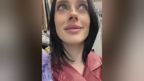 Video of a smiling woman with straight black hair and a red leather jacket, looking up with light blue eyes, in a cluttered room with hanging clothes and a purple garment in the background.