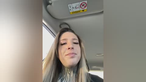 Media: Video of a young woman with long blonde hair and fair skin, wearing a striped turtleneck, smiling from the driver's seat of a car.