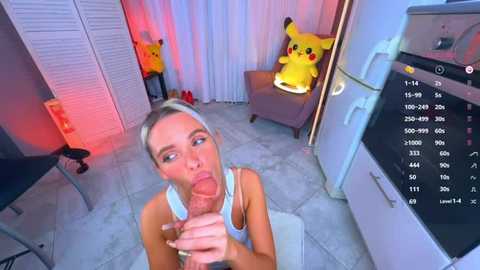 Video of a blonde woman with fair skin, wearing a white tank top, kneeling on a tiled floor, giving a blowjob to a man's erect penis. Background features a Pikachu plush, a TV, and a red lamp.