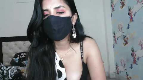 Media: Video of a woman with long black hair, wearing a black mask, black top, and floral-patterned wallpaper in a bedroom.