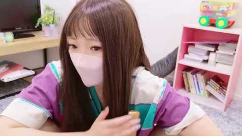 Video of an Asian girl with long brown hair, wearing a white mask, purple and white-striped shirt, and holding a cup, in a tidy room with shelves, books, and a plant.