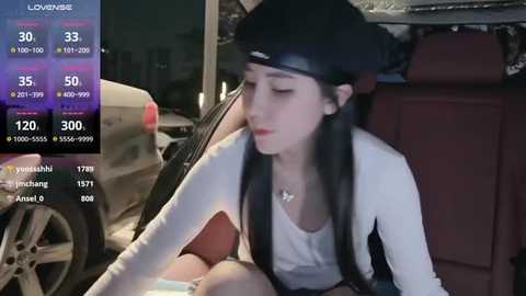 Media: A video of a young woman with long black hair, wearing a black beanie, white top, and black shorts, sitting in a car, looking at a phone screen displaying streaming metrics.