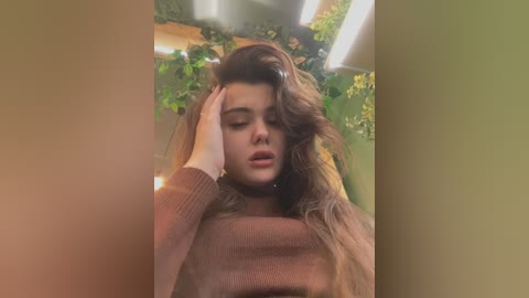 Media: A video captures a young woman with long, wavy brown hair, wearing a brown sweater, in an indoor greenhouse setting. She has her hand on her forehead, appearing thoughtful or tired.