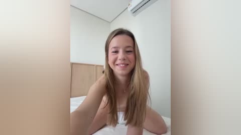 Media: Video of a smiling, nude young woman with long blonde hair, seated on a bed in a minimalist bedroom with white walls and light wooden furniture.