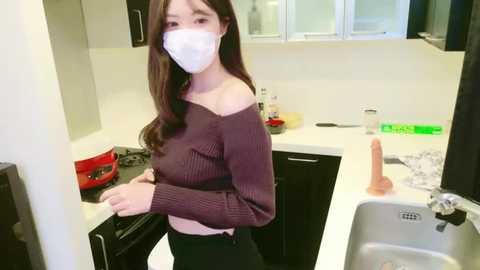 Media: Video of an Asian woman with long brown hair, wearing a white mask, a brown off-shoulder sweater, and black pants, standing in a modern, cluttered kitchen with white cabinets, a sink, and a red bowl.