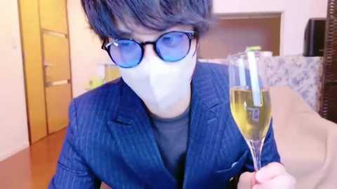 Media: Video of a man with short, dark hair wearing blue pinstripe suit, white mask, and blue-tinted glasses, holding a champagne flute in a modern, brightly-lit room.