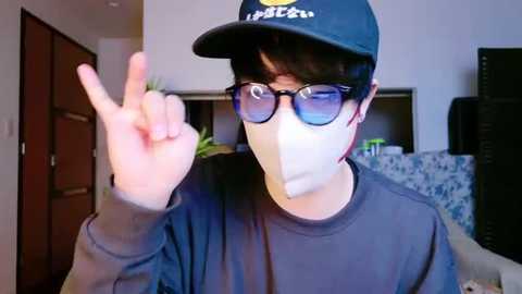 Media: Video of a young person with short black hair, wearing a black cap, blue-rimmed glasses, and a mask, making a peace sign with their right hand, in a modern living room with wooden doors and a blue-patterned couch.