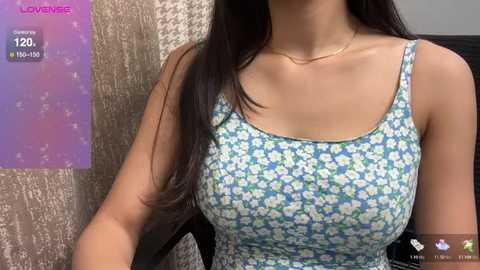 Media: Video of a woman with long black hair, wearing a blue floral tank top, sitting in a room with a beige textured wall and a purple digital clock displaying 12:20.