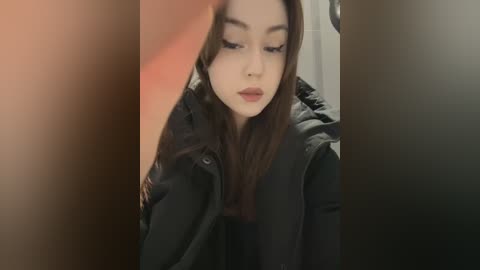 A video of a young woman with long, straight brown hair, fair skin, and dark eyes, wearing a black puffy jacket, taken indoors with soft lighting.