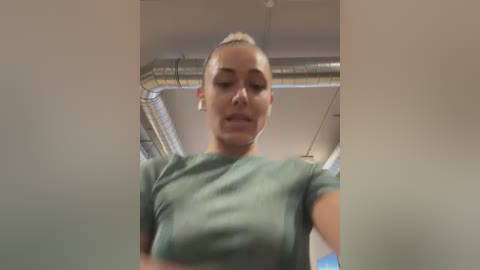 Media: Video of a fit woman with blonde hair in a ponytail, wearing a green t-shirt, taken from a low angle in an industrial setting with visible pipes and ducts in the background.