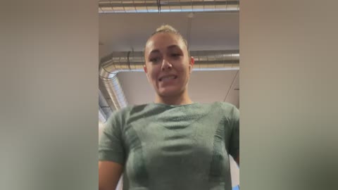 Media: Video of a smiling, light-skinned woman with short, blonde hair, wearing a teal, short-sleeved shirt. Background shows a modern, industrial ceiling with metal pipes and white walls.