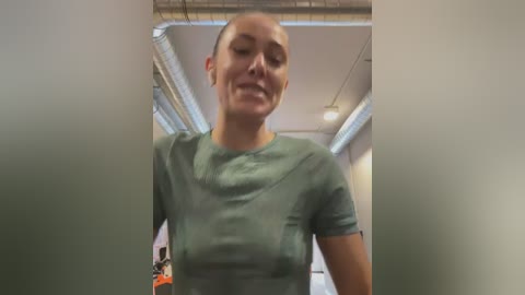 Media: Video of a bald Caucasian woman in a green t-shirt, slightly blurred, standing in a dimly lit, industrial room with exposed pipes and a ceiling fan.