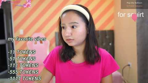 Media: Video of an Asian woman with straight black hair, wearing a pink shirt, standing indoors. Background features a striped wall and pink furniture. Text overlays mention \"my favorite videos\" and \"tokens.\