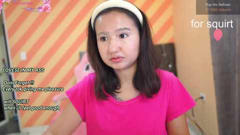 Media: A video of an Asian woman with shoulder-length black hair, wearing a pink t-shirt and headband, standing in a room with striped orange walls and pink heart decorations.