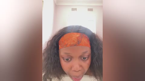 A video of a Black woman with medium brown skin and curly black hair, wearing an orange headband, sticking her tongue out, set in a small, brightly lit bathroom with white walls and a door.