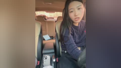 Video of an Asian woman with long black hair, wearing a dark blue sweater, sitting in the back seat of a van. The van's interior is beige with a tablet on a seat.