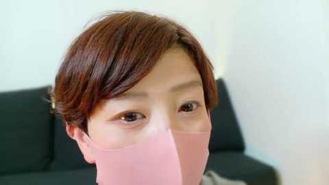 Media: Video of an Asian woman with short, dark brown hair, wearing a pink face mask, gazing intently at the camera. Background features a black sofa.