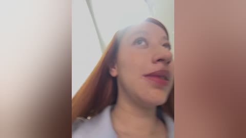 A blurred video of a young woman with long red hair, wearing a light-colored shirt, looking upward with a slight smile. The background is out of focus, suggesting an indoor setting.