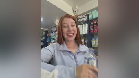 Video of a smiling young Caucasian woman with red hair in a pharmacy, wearing a light blue zip-up jacket, holding a clear plastic bottle of liquid. Background shows shelves of medical supplies and products.