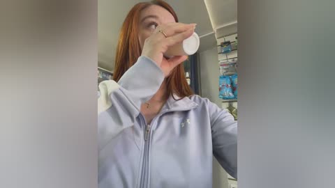 Video of a young, fair-skinned woman with long red hair, wearing a light blue hoodie, drinking from a white plastic bottle in a pharmacy with shelves filled with products in the background.