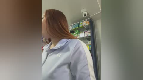 Video of a woman with long auburn hair in a light blue zip-up jacket, standing in a brightly lit, clean room with a surveillance camera overhead.