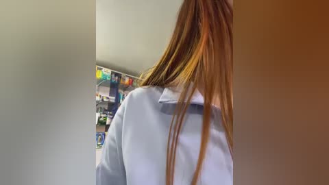 A video of a woman's back, with long, straight brown hair, wearing a light blue polo shirt, standing in a well-lit room with a shelved wall of colorful products.