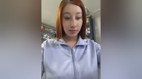 A video of a young Asian woman with straight, shoulder-length auburn hair, wearing a light blue zip-up jacket, standing in a brightly lit room with shelves of items in the background.