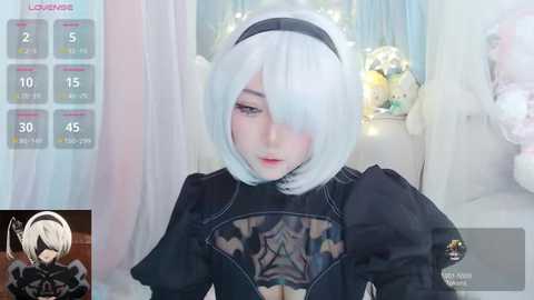 Media: Video of a pale-skinned woman with short white hair, wearing a black dress and black hairband, in a soft, pastel room with a plush toy, a calendar, and a game UI overlay.