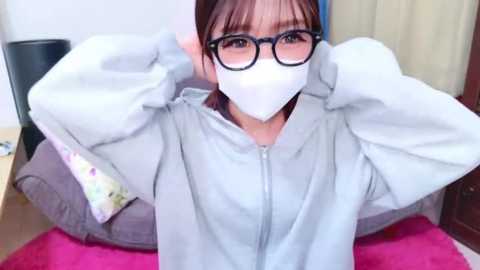 Media: Video of an East Asian woman with fair skin, dark hair, and large glasses, wearing a white mask and a light gray hoodie, sitting on a pink blanket, indoors.