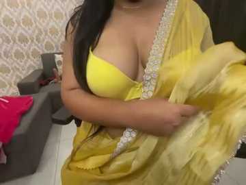 Media: Video of a woman in a yellow saree with a deep cleavage, holding the fabric. Background shows a grey sofa with a pink pillow and patterned wallpaper.