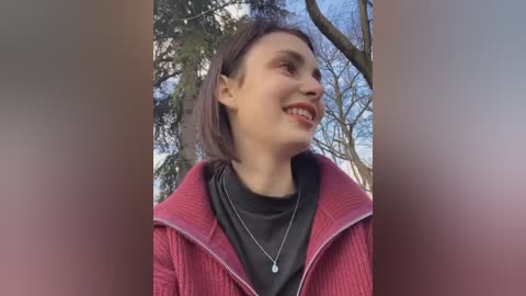 Media: Video of a smiling woman with fair skin, brown hair, and red lipstick, wearing a black turtleneck and a red zip-up jacket, set against a background of bare trees and a blue sky.