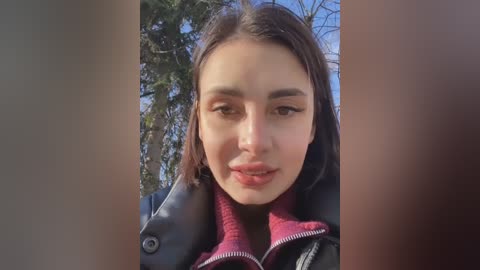 Media: Video of a young woman with light skin and dark hair, wearing a black jacket with a red lining, standing outdoors near trees. She has a neutral expression.