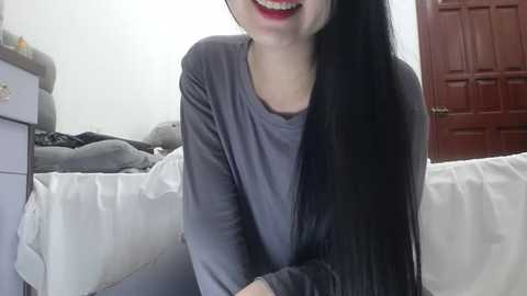 Media: Video of a smiling Asian woman with long black hair, wearing a gray sweater, seated on a white bed in a dimly lit room with a red door in the background.