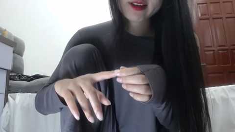 Media: Video of a woman with long black hair, wearing a grey long-sleeve top, seated on a bed with white sheets. She has fair skin and is smiling, pointing with her right hand. Background includes a white wall and a closed brown door.