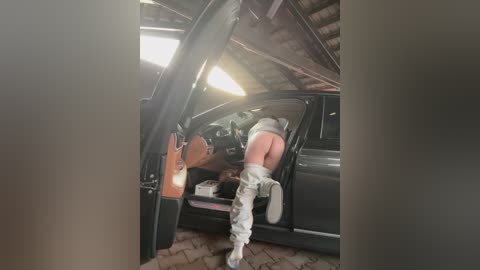 Media: Video of a woman with pale skin, wearing white leggings and boots, partially exiting a black SUV, in a dimly lit garage with wooden beams.