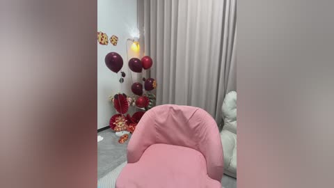 Media: Video of a modern room with a plush pink armchair, adorned with red heart balloons and floral arrangements on a white wall. Beige curtains and a cozy white couch complete the scene.