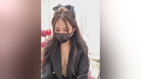 Video of an East Asian woman with long black hair, wearing a black face mask, black jacket, and black headband, indoors with white walls and red decorative balls in the background.