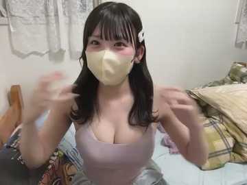 Media: A video of an Asian woman with long black hair and a white hairclip, wearing a beige mask and a low-cut light pink top, standing in a messy bedroom with white curtains, a plaid blanket, and a bed.