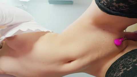 Video of a light-skinned woman's torso, wearing black lace stockings, with a pink vibrator inserted into her anus. The background is blurred, featuring a white wall.