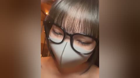 Media: Video of a person with straight, dark hair and bangs, wearing black-rimmed glasses and a white face mask, set against a warm, blurred background.