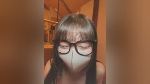 Media: Video of a woman with long black hair, wearing black-rimmed glasses and a white face mask, indoors with warm lighting, blurry background.