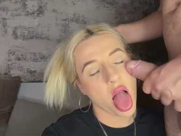 A video shows a blonde woman with short hair, wearing a black shirt, kneeling with eyes closed, tongue out, and a man's erect penis near her face.