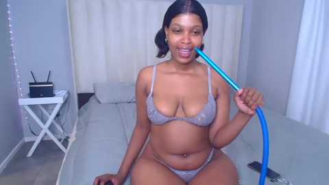 Video of a young Black woman with medium skin tone and dark hair in a ponytail, wearing light gray lace lingerie, blowing a blue straw into her mouth while sitting on a white bed in a minimalistic bedroom with white curtains and a white nightstand.