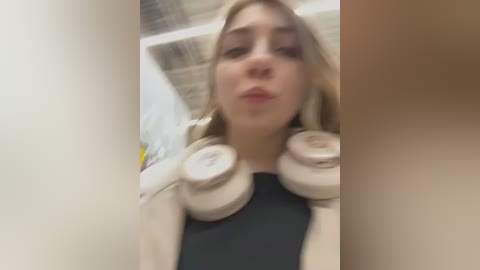 Media: A video of a young woman with blonde hair and pale skin wearing beige headphones and a black top, taken in a brightly lit room with white walls and a metal ceiling.