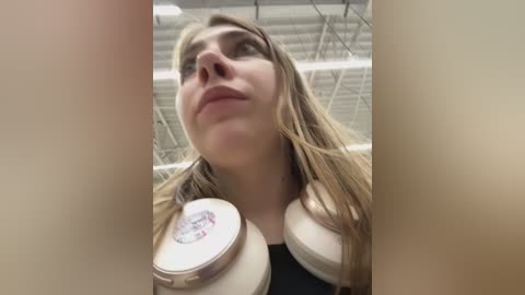 Media: Video of a young Caucasian woman with long blonde hair, wearing a black top, looking up with a confused expression. She's holding large white headphones in front of her face. Background shows a gym with industrial ceiling lights.