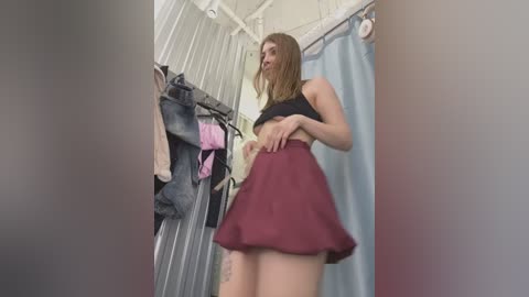 Media: Video of a young woman in a fitted black crop top and maroon skirt, standing in a cluttered closet with hanging clothes and a blue curtain in the background.