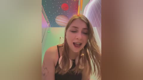 Media: A video of a young woman with long blonde hair, wearing a black tank top, leaning against a wall with colorful space-themed decals. She appears joyful, with a slight smile.