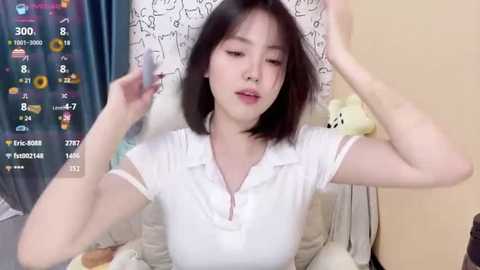 Media: Video of an Asian woman with straight, shoulder-length black hair, wearing a white polo shirt, sitting on a bed. Background features a teddy bear and a patterned pillow.