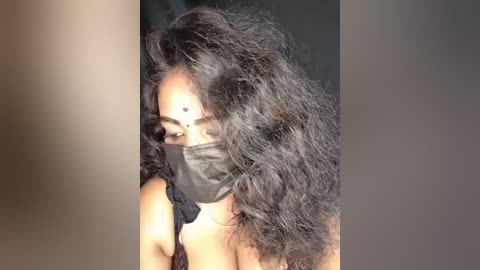 Media: Video of a woman with long, voluminous black curly hair partially covering her face, wearing a black mask and a red bindi on her forehead.